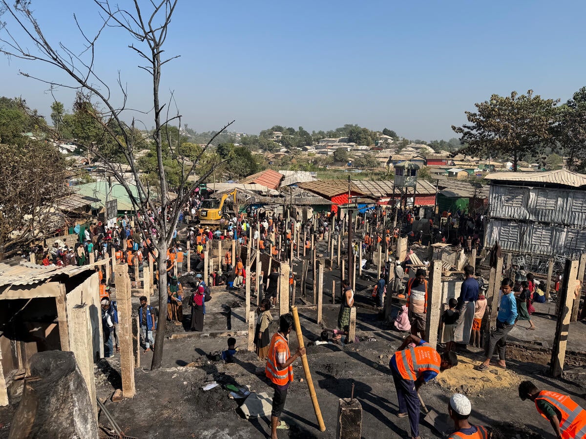 Scene of a fire devastation in a refugee camp where aid workers erect wooden beams to begin restoration efforts.