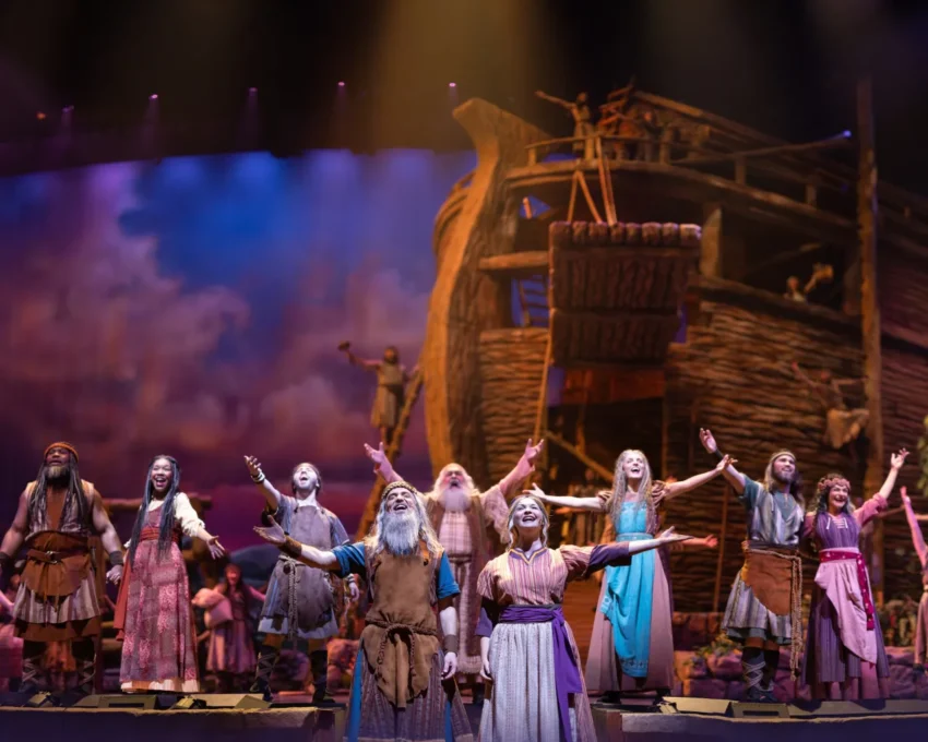 Actors in colorful costumes perform before a large wooden ark on stage