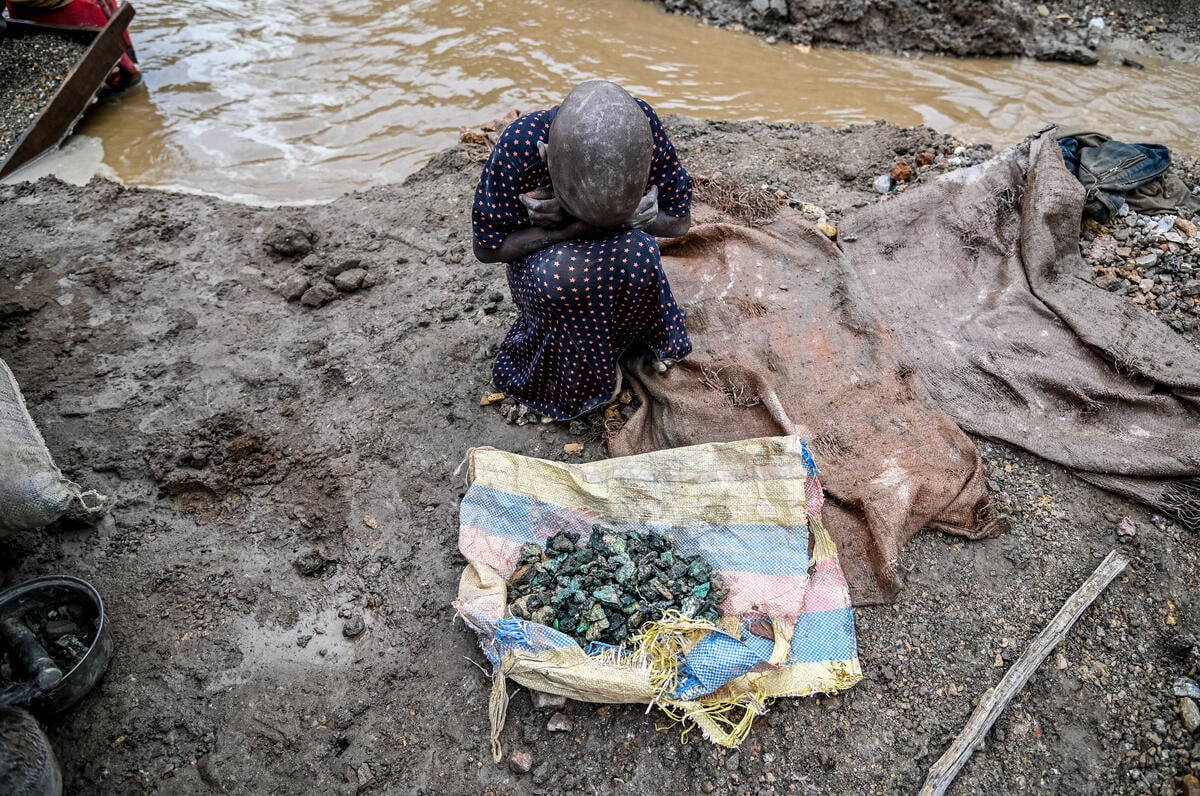 A young girl sits curled up with her head down in front of a pile of sorted materials and rocks on the edge of mud and water.