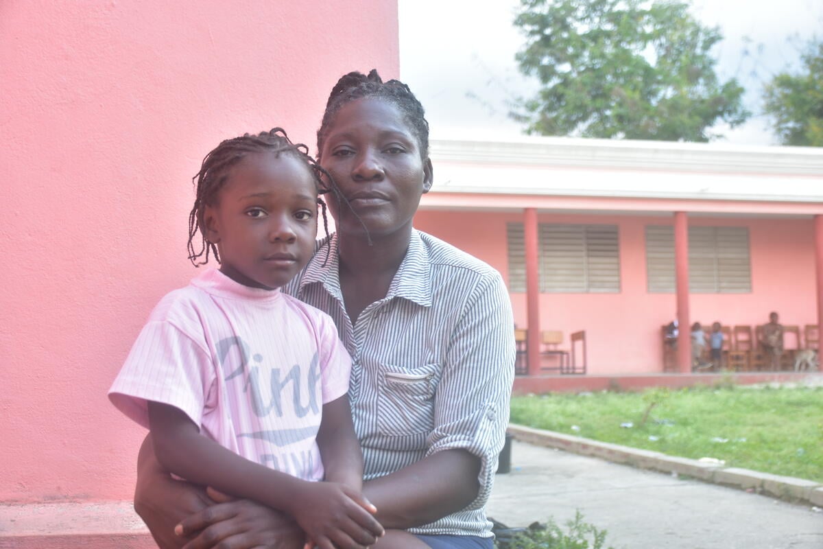 A young girl in a pink shirt sits on a woman’s lap, both gazing at the camera in front of a pink building.