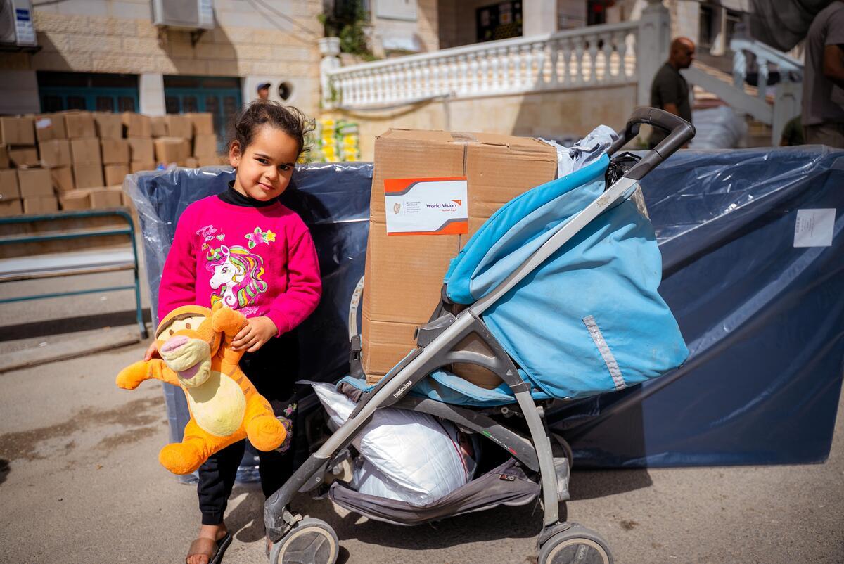 A young girl holding a Tigger stuffed toy stands near a stroller and boxes, with a house and supplies in the background.