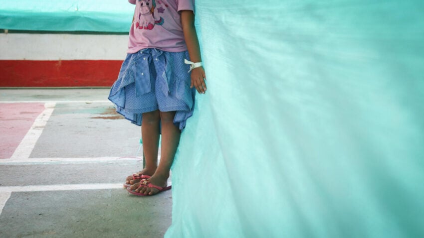 A young girl stands against a tarp in a temporary shelter.