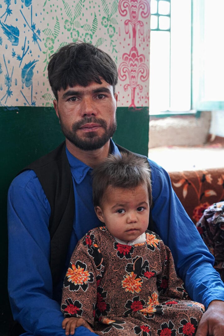 A man with a dark beard and mustache looks into the camera as a young girl in a floral dress sits on his lap.