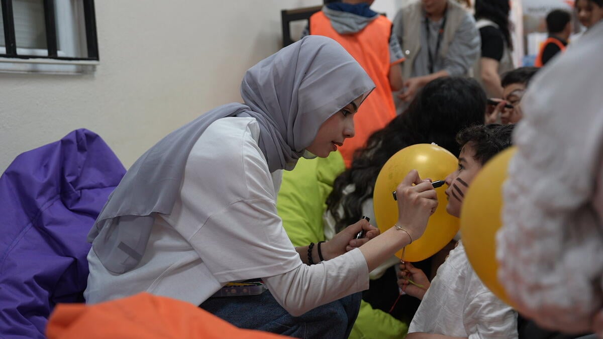 A girl leans forward from her chair to draw on a young boy’s face, surrounded by balloons and people. 