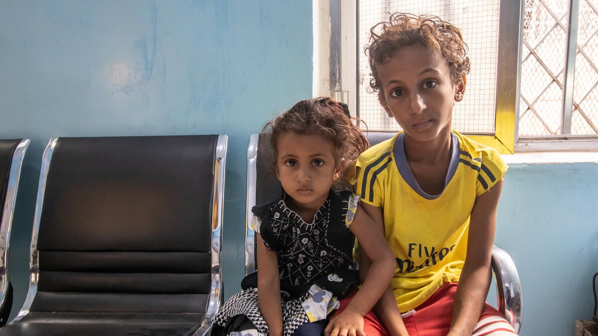 Two young children with visible signs of malnutrition sit closely together in a chair in a blue-walled room. 