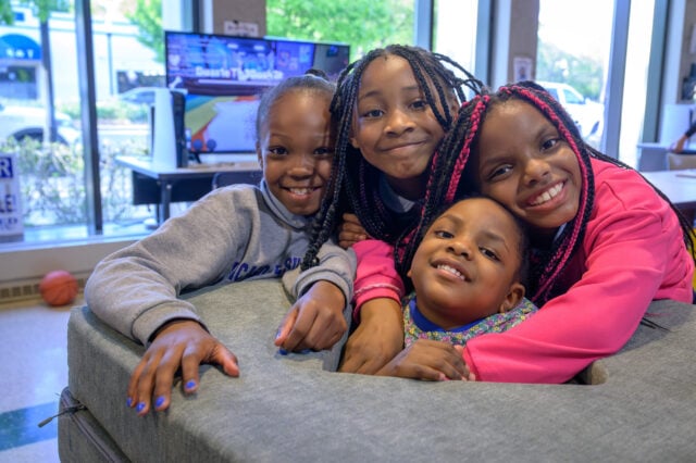 Four children smile looking at the camera. They crowd around a pile of cushions. Behind them is a TV screen.