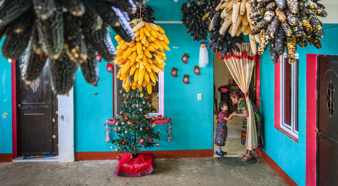 A woman talks to a child next to a Christmas tree, below a ceiling hung with drying bunches of maize corn.