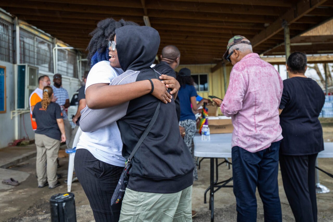 Two people hug outdoors as others stand nearby under a shelter with tables.