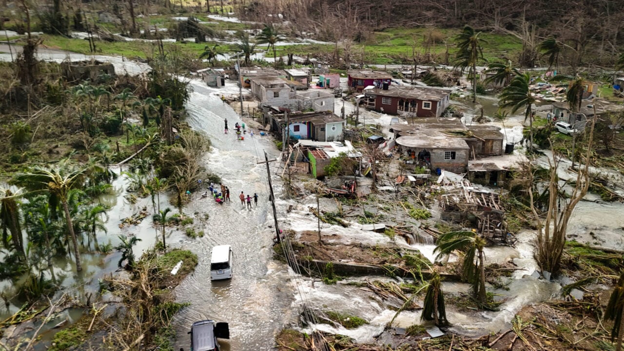 Two vans try driving through a flooded street. Downed trees, damaged homes, and people navigating a flooded street are visible from a bird’s-eye’s view of a community after a hurricane.