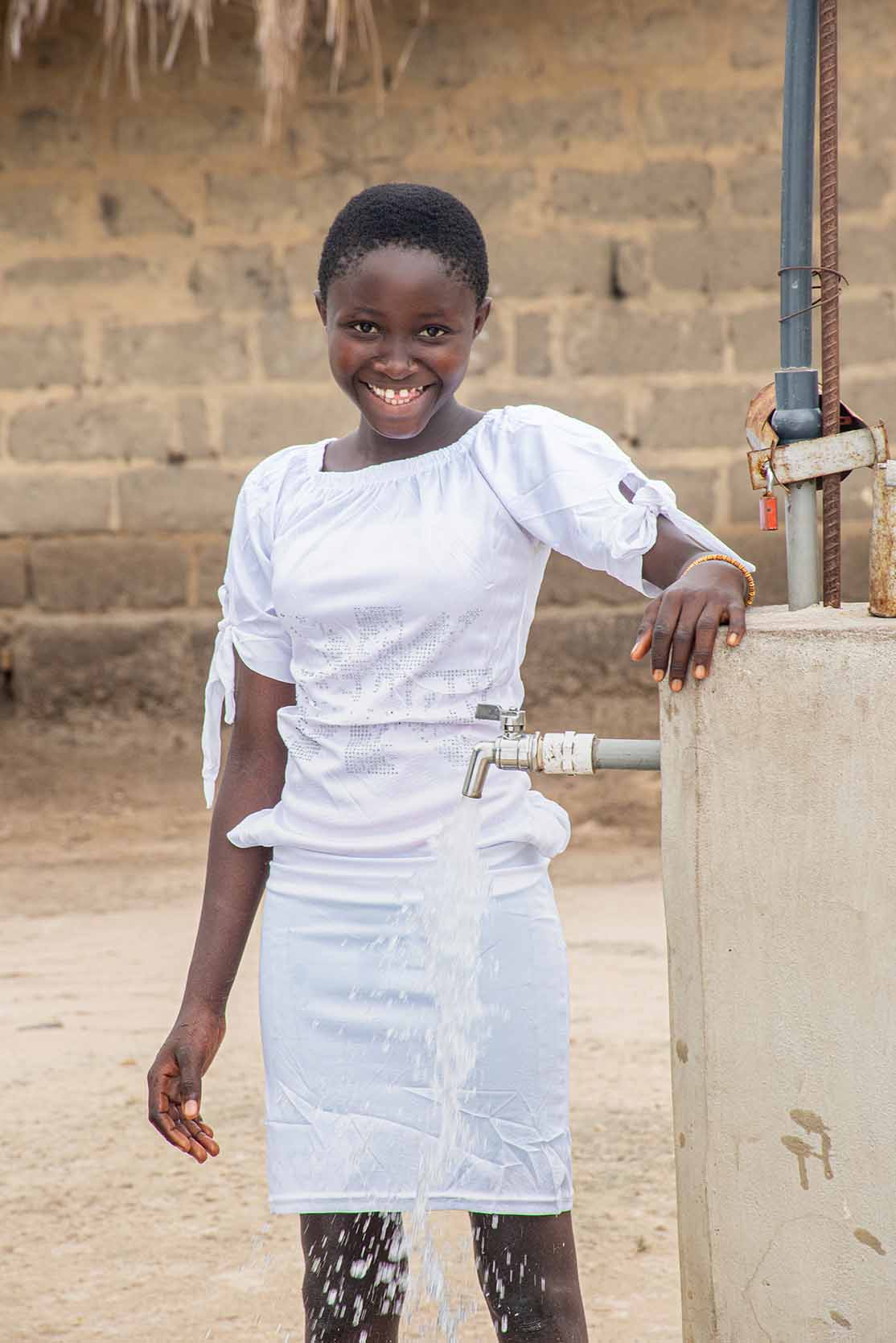 A young girl stands behind a water tap, smiling widely as water splashes out.