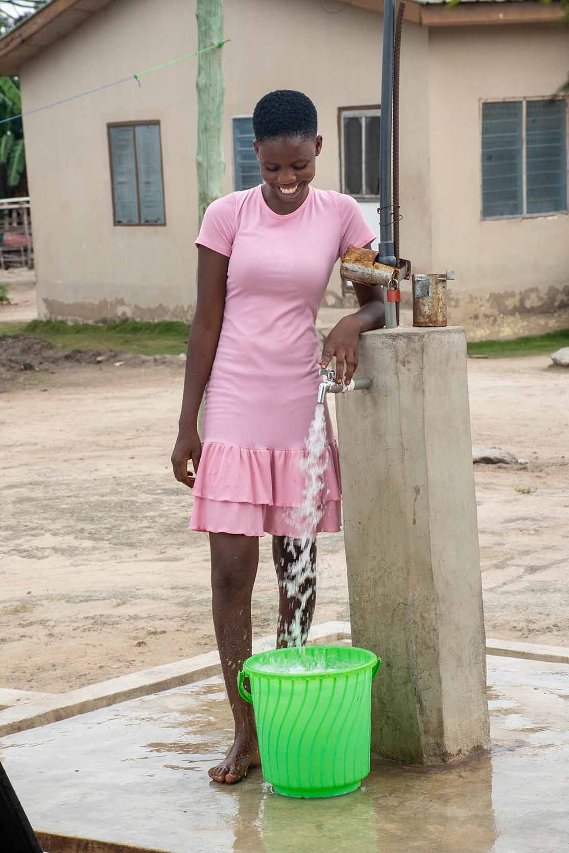 A young woman smiles while watching water pour from the tap into her bucket.