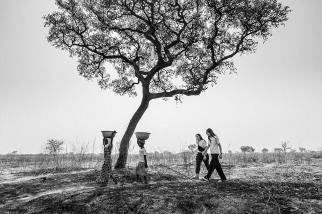 A black-and-white photograph of two young African girls walking underneath a tree, carrying basins of water on their heads, followed by two American girls.