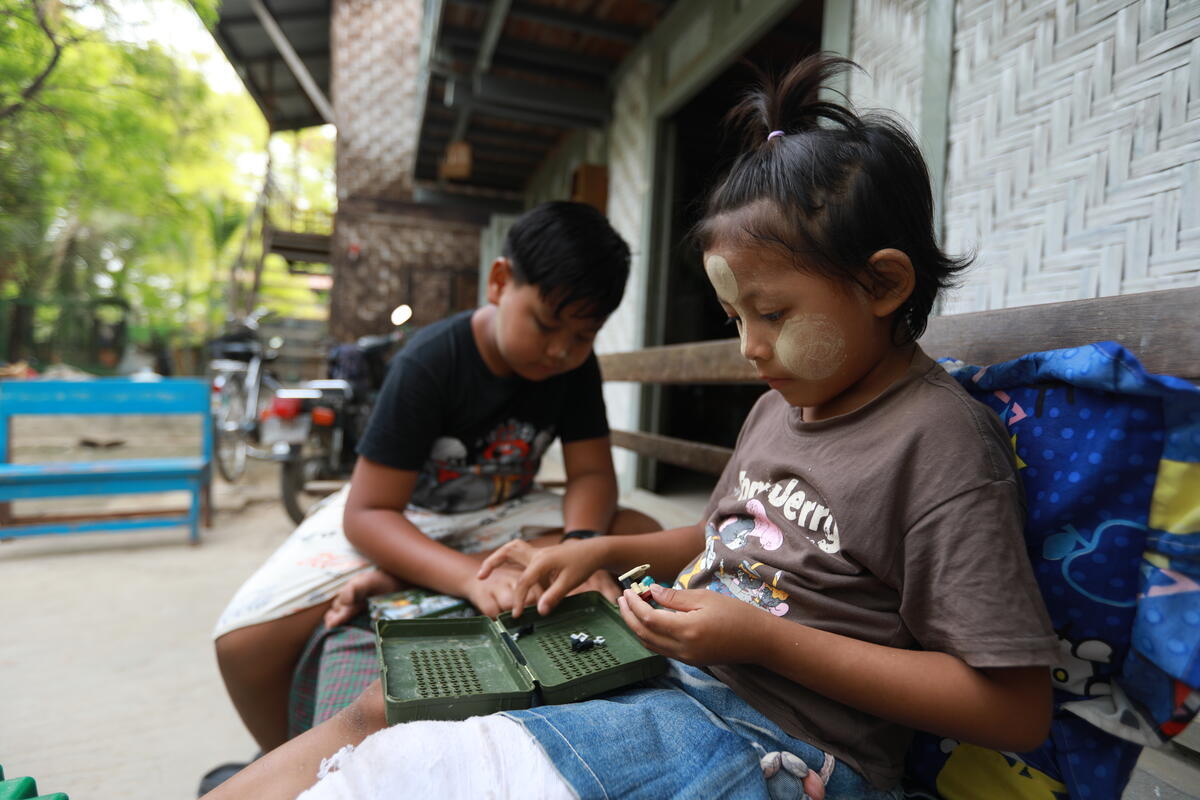 Two children sit on a bench; one girl wears traditional protective paste, thanaka, on her cheeks and forehead, and her leg is in a white cast. 