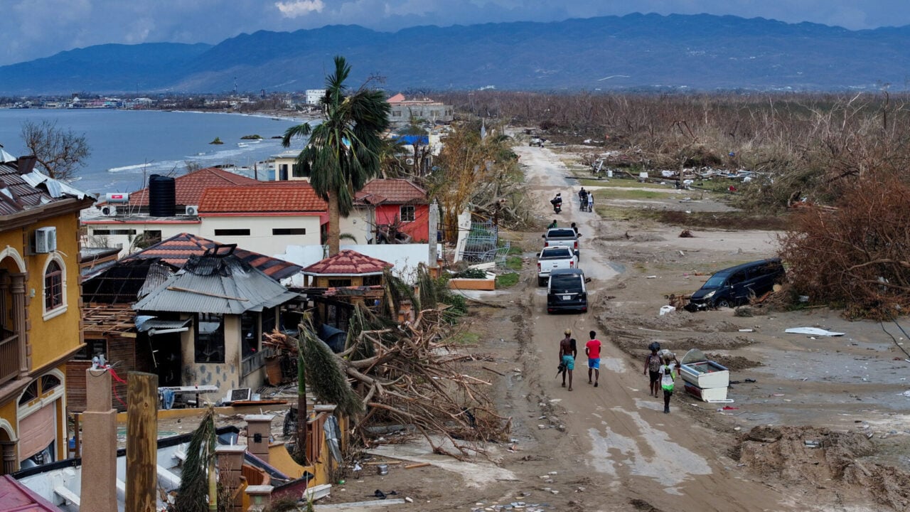 An aerial view of collapsed seaside homes with debris and aid personnel on a dirt road after a deadly hurricane in Jamaica.