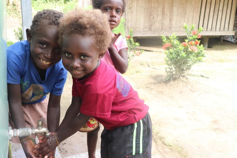 Two smiling children wash their hands at an outdoor tap, and another child stands behind.