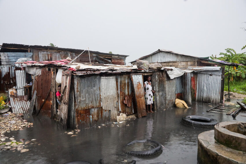 A woman stands in the doorway of a tin-sheet home surrounded by floodwaters, while another person peeks out from an opening.