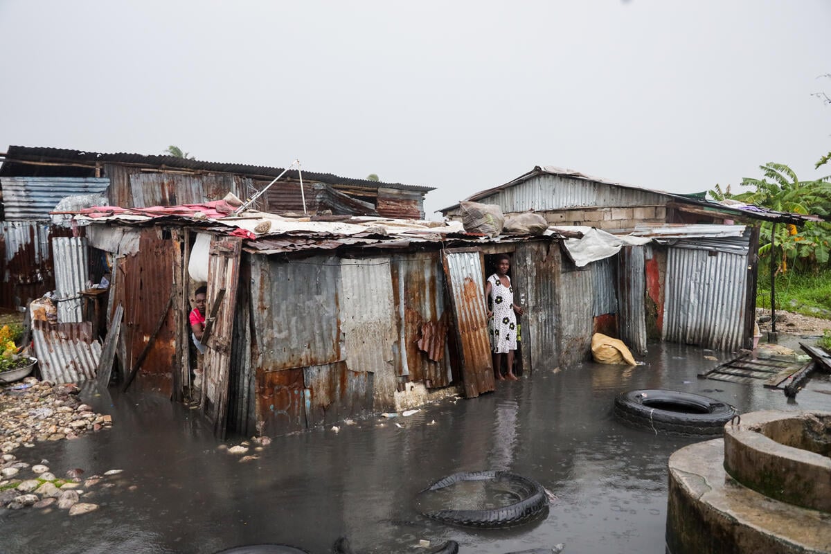 A woman stands in the doorway of a tin-sheet home surrounded by floodwaters, while another person peeks out from an opening.