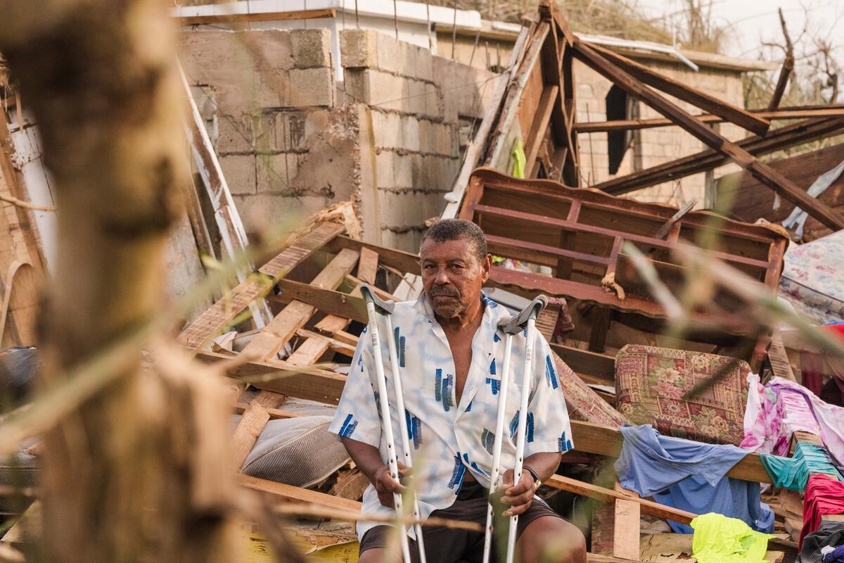 A man with a mustache holds crutches while sitting among the ruins of his storm-damaged home. 