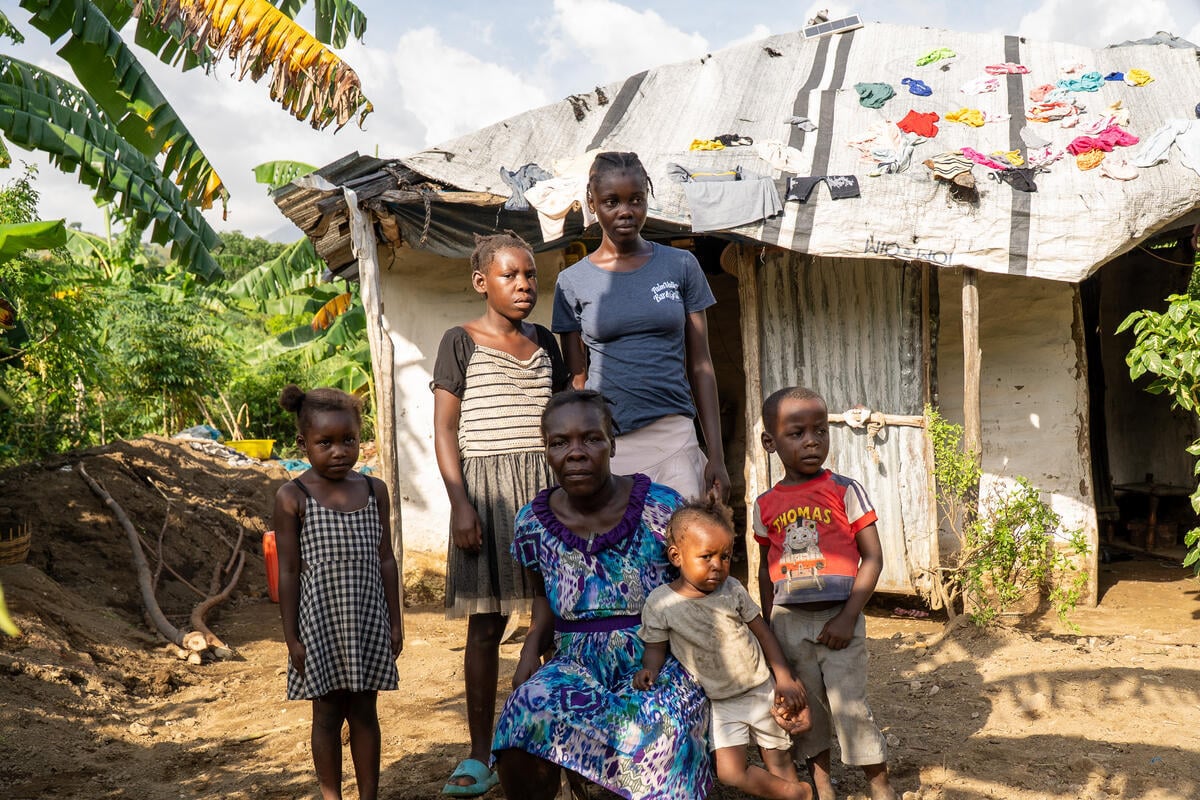 A mother surrounded by children sits outside a damaged home. The tin roof is partially covered by a tarp with colorful clothes set out to dry. 