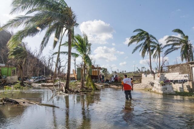 A man in a World Vision vest wades through calf-deep floodwater toward residents waiting by a damaged home. Palm trees lean in the storm’s aftermath.