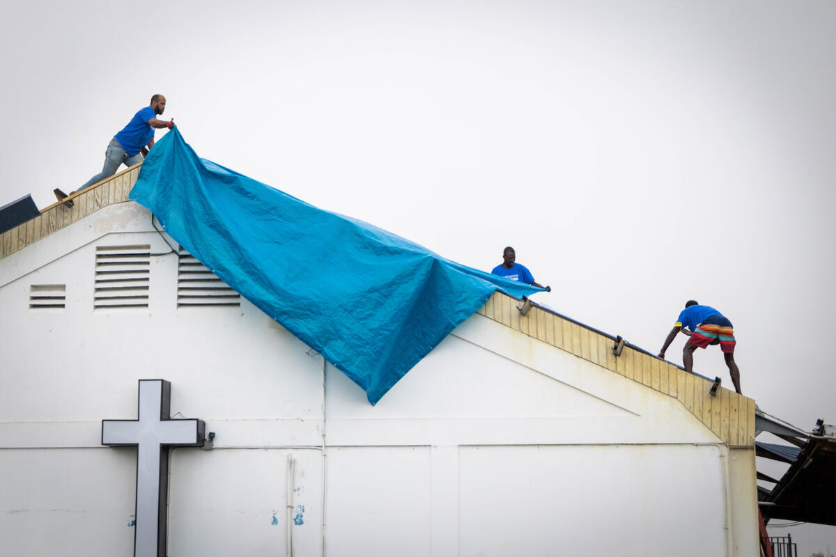 Three men wearing matching T-shirts secure a tarp on the roof of a church building damaged by a hurricane. A cross is visible on the wall below them.