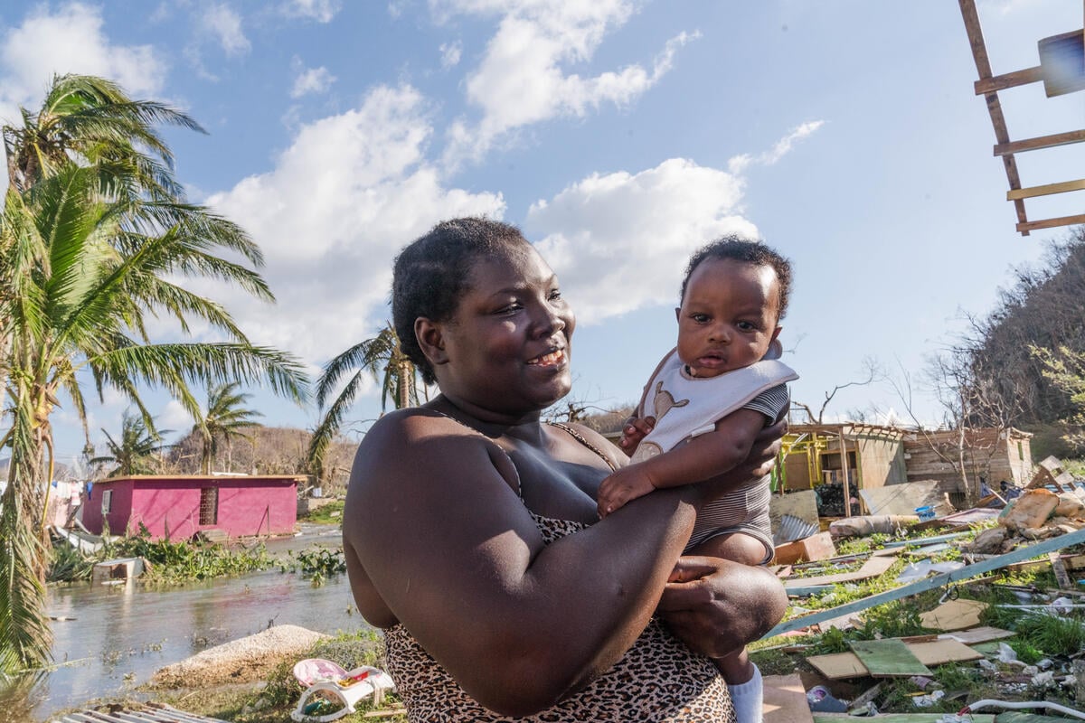 A woman holds a baby boy surrounded by debris and flooded ground in Jamaica after Hurricane Melissa. A leaning palm tree and gray clouds fill the background.