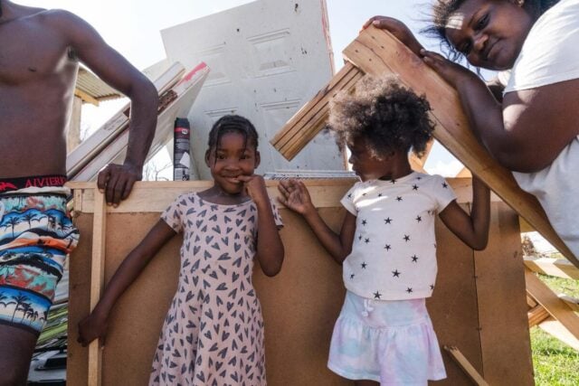 Two young girls stand and smile between two adults, all in front of wooden debris.