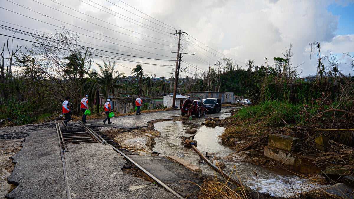Four aid workers carrying supplies walk in a line along a flooded, damaged road covered with debris and downed vegetation. Two men are visible near each of the two vehicles parked at the scene.
