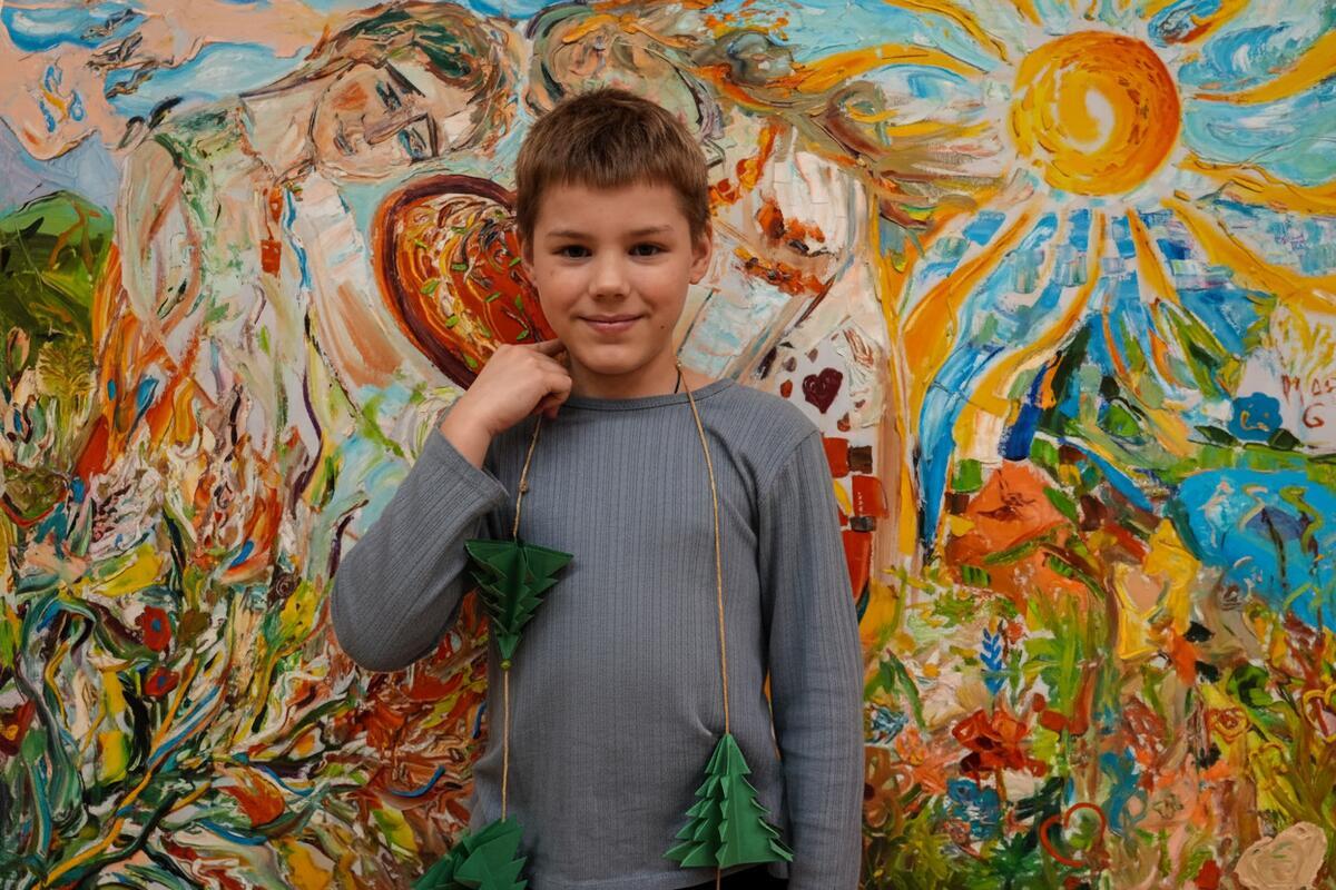 A smiling boy stands before a colorful painting, holding a string of paper Christmas trees around his neck.