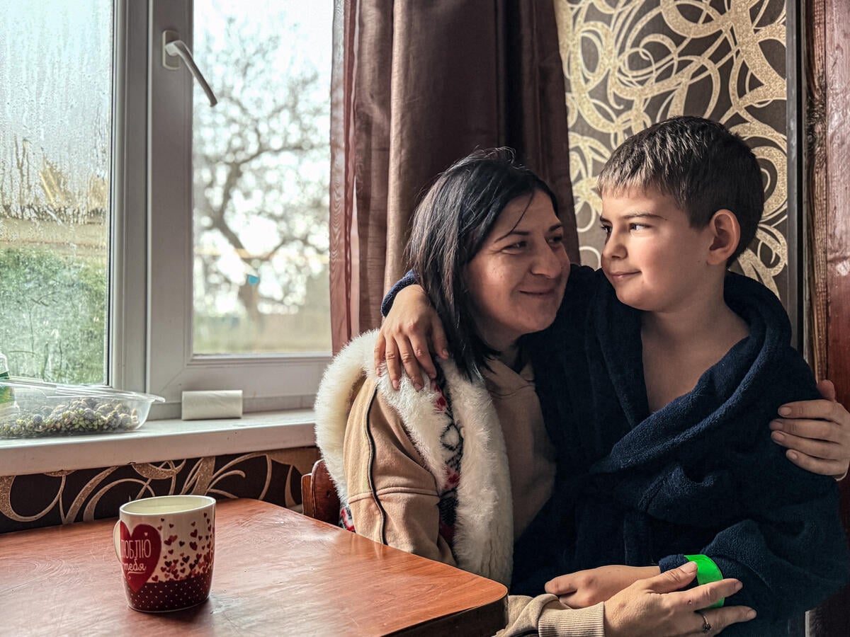 A mother with dark hair sits indoors with her son, holding him close as they look at each other near a window.