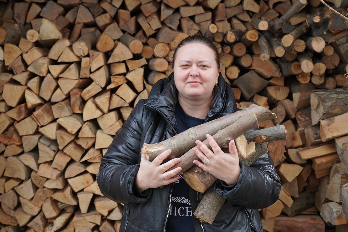 A woman in a black jacket holds firewood in her arms while standing in front of a stacked woodpile, highlighting efforts to stay warm during the winter in Ukraine.