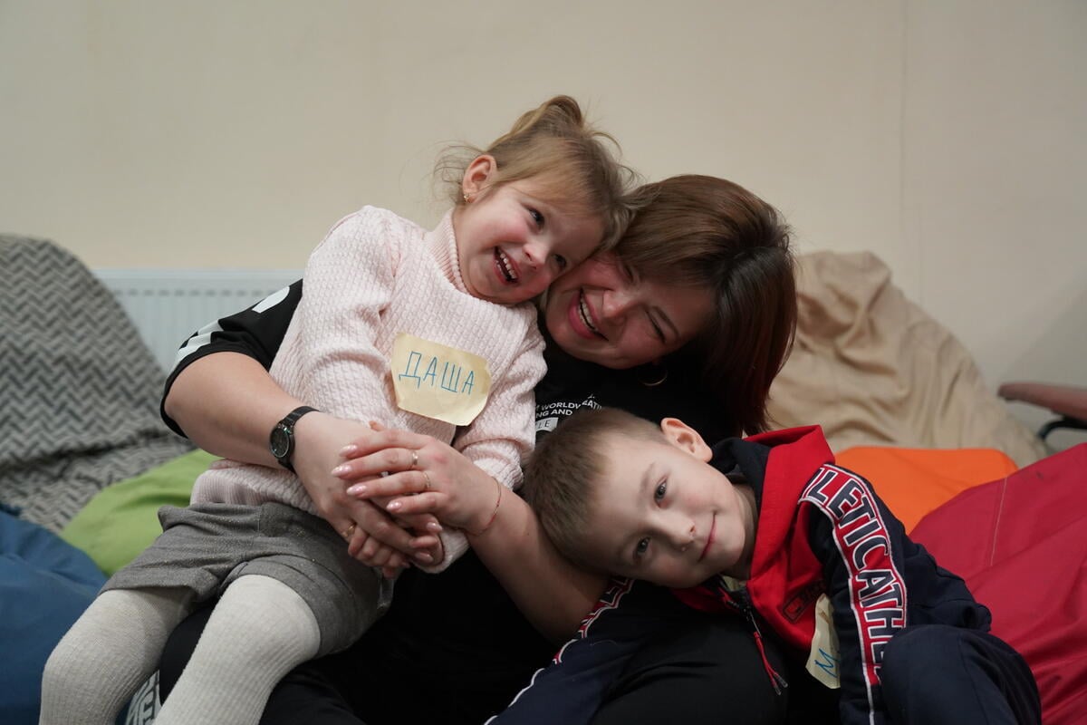 A mother and her two young children surrounded by pillows are seen laughing and hugging each other.