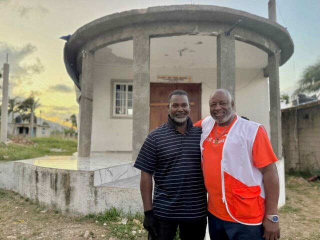 Two men smile for a photo in front of a building. The sun is setting behind them.