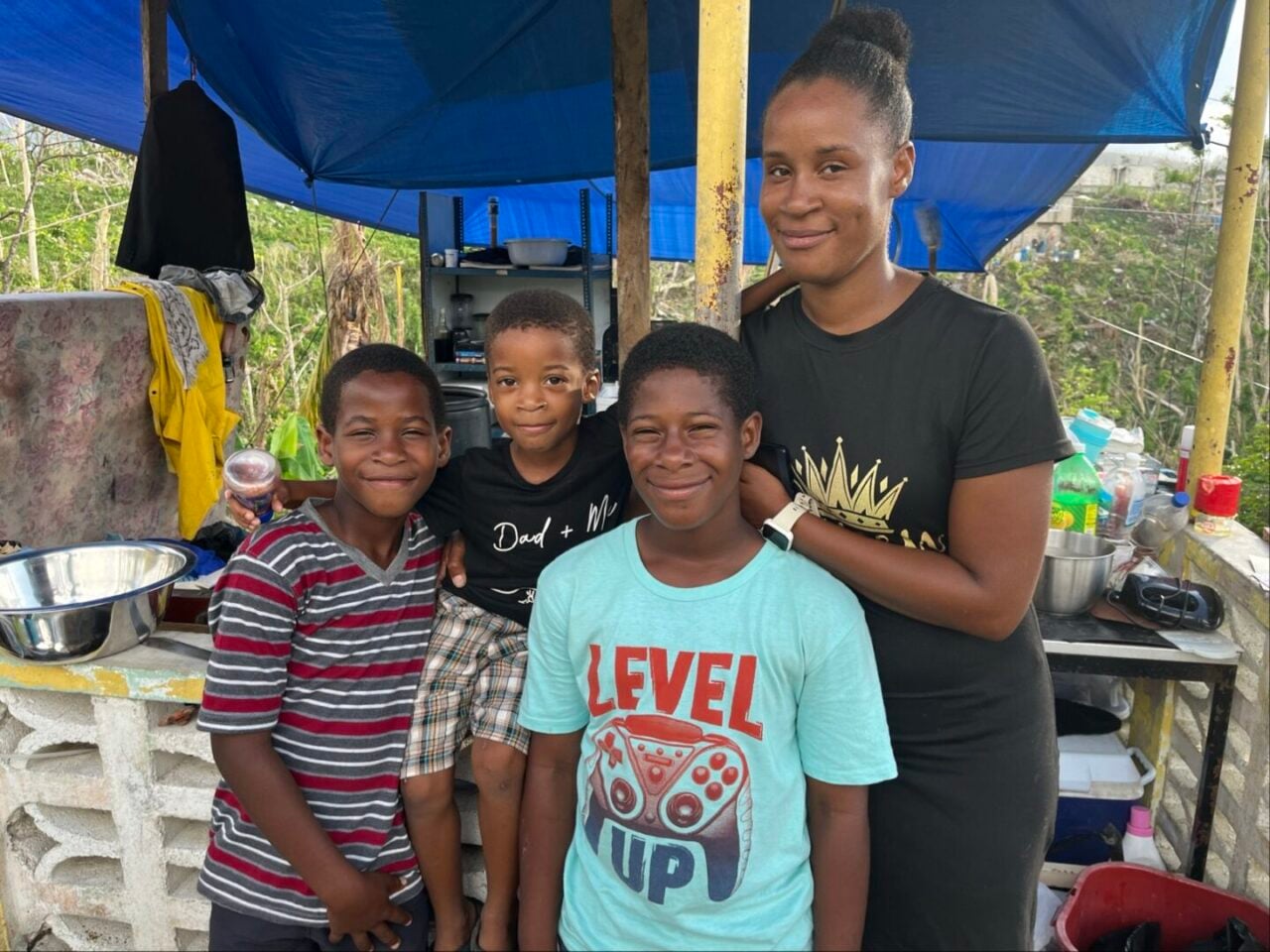 A woman stands under a tarped shelter with three boys.