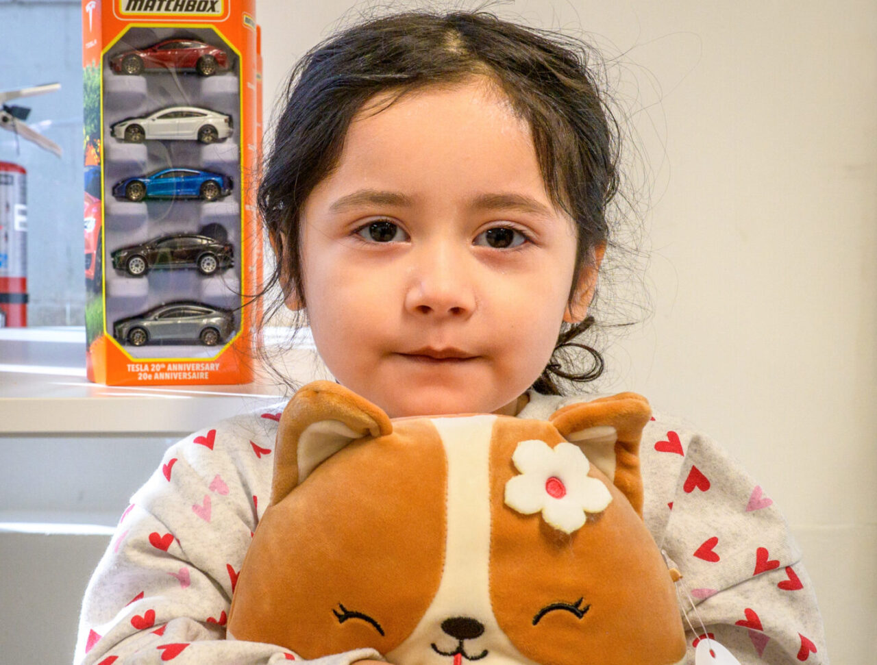 A little girl hugs a plush toy to her chest. On a table behind her stands a box of miniature cars.