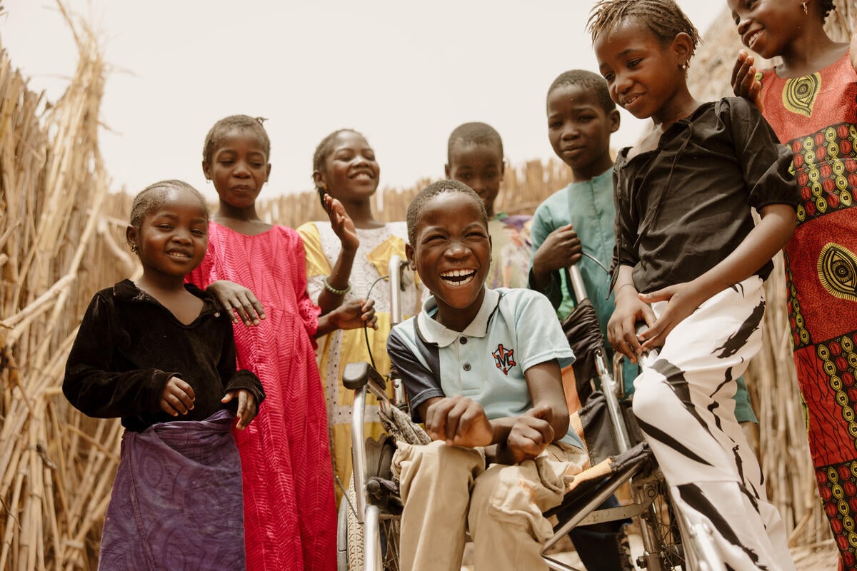 A boy in a wheelchair beams with a wide smile as he is surrounded by other children, with tall, dry stalks in the background.