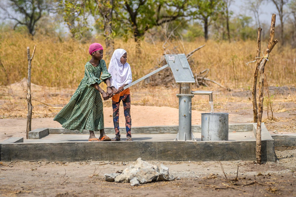 Two girls with headscarves stand at a water pump, filling a large metal container with clean water. 