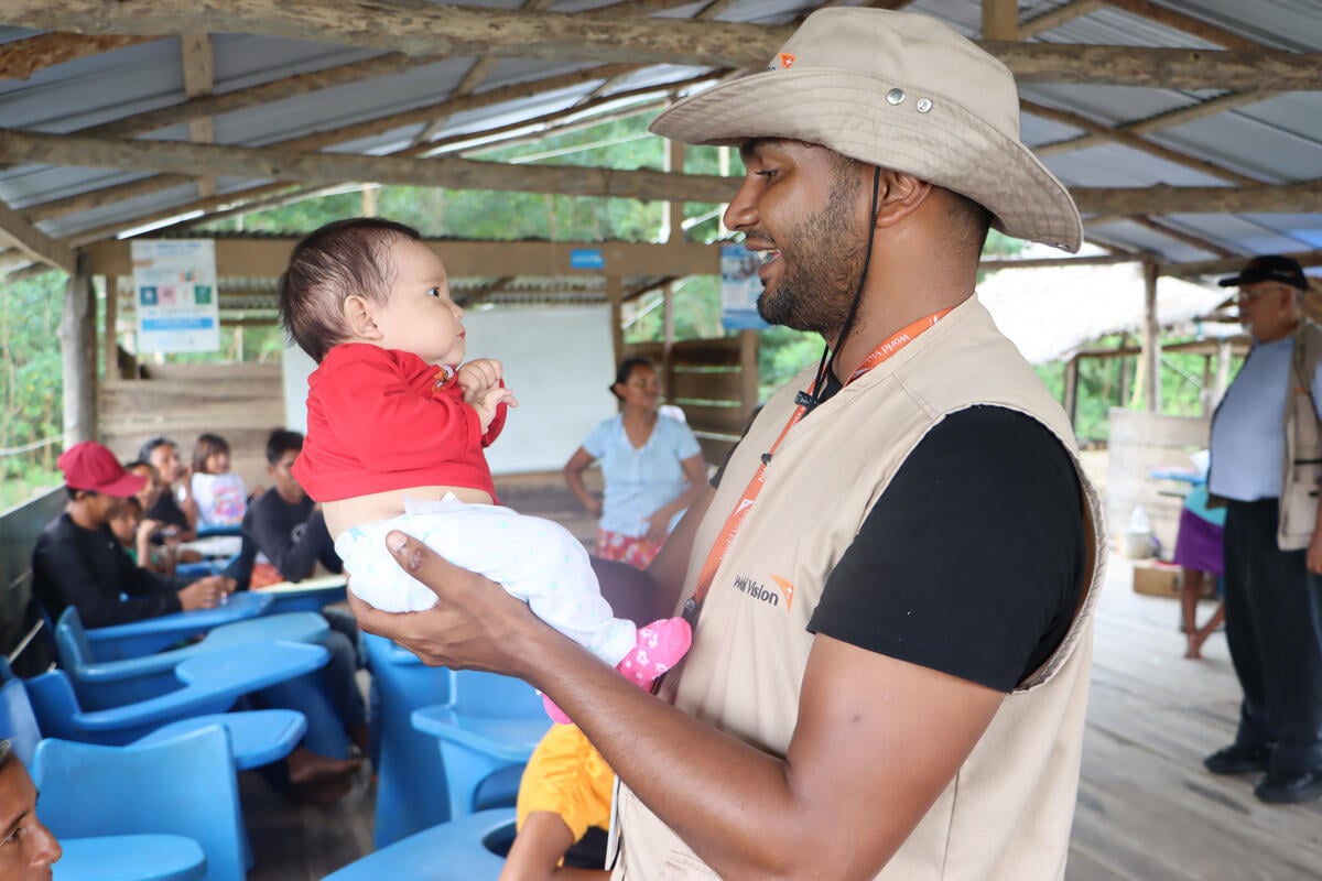 An aid worker wearing a hat and vest smiles while holding a baby inside a community meeting space. 