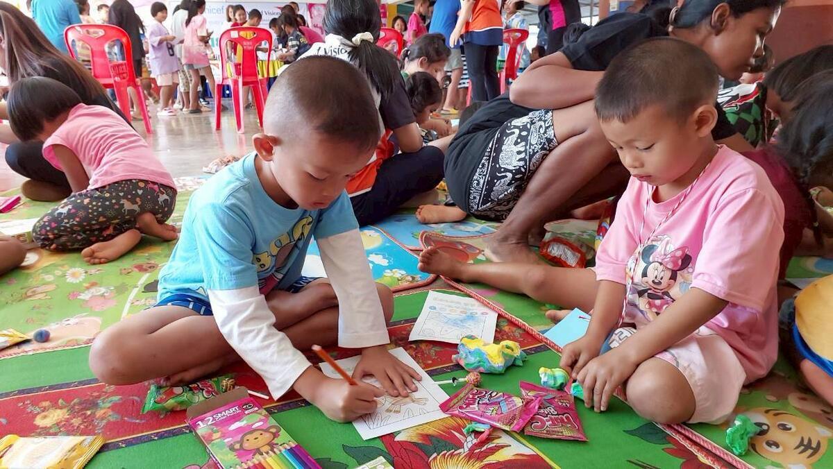 A ground-level view of children sitting on colorful mats on the floor, drawing and playing in a bright room filled with other children. 