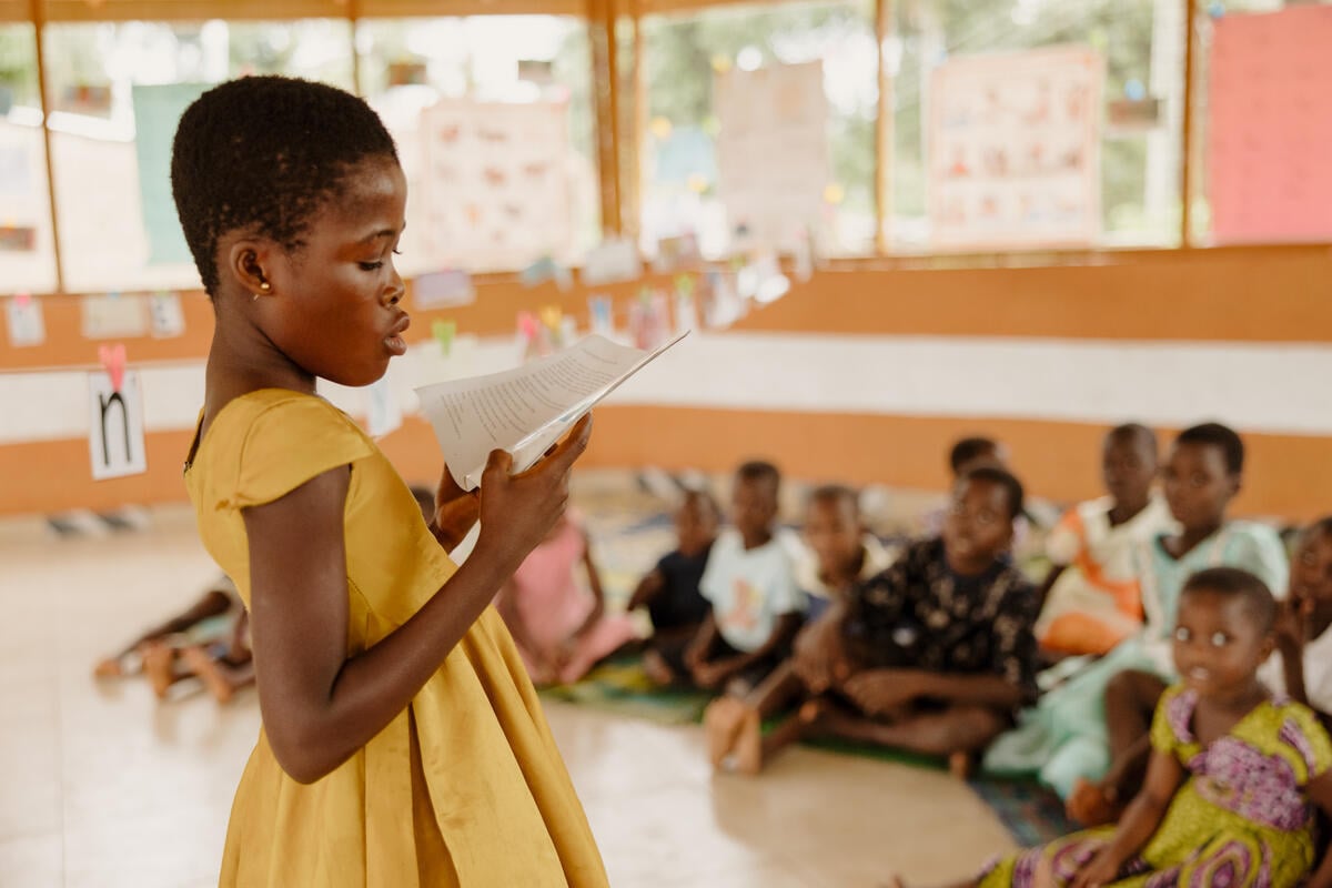 A girl reads aloud from a book in front of a group of children sitting on the floor of a bright classroom.