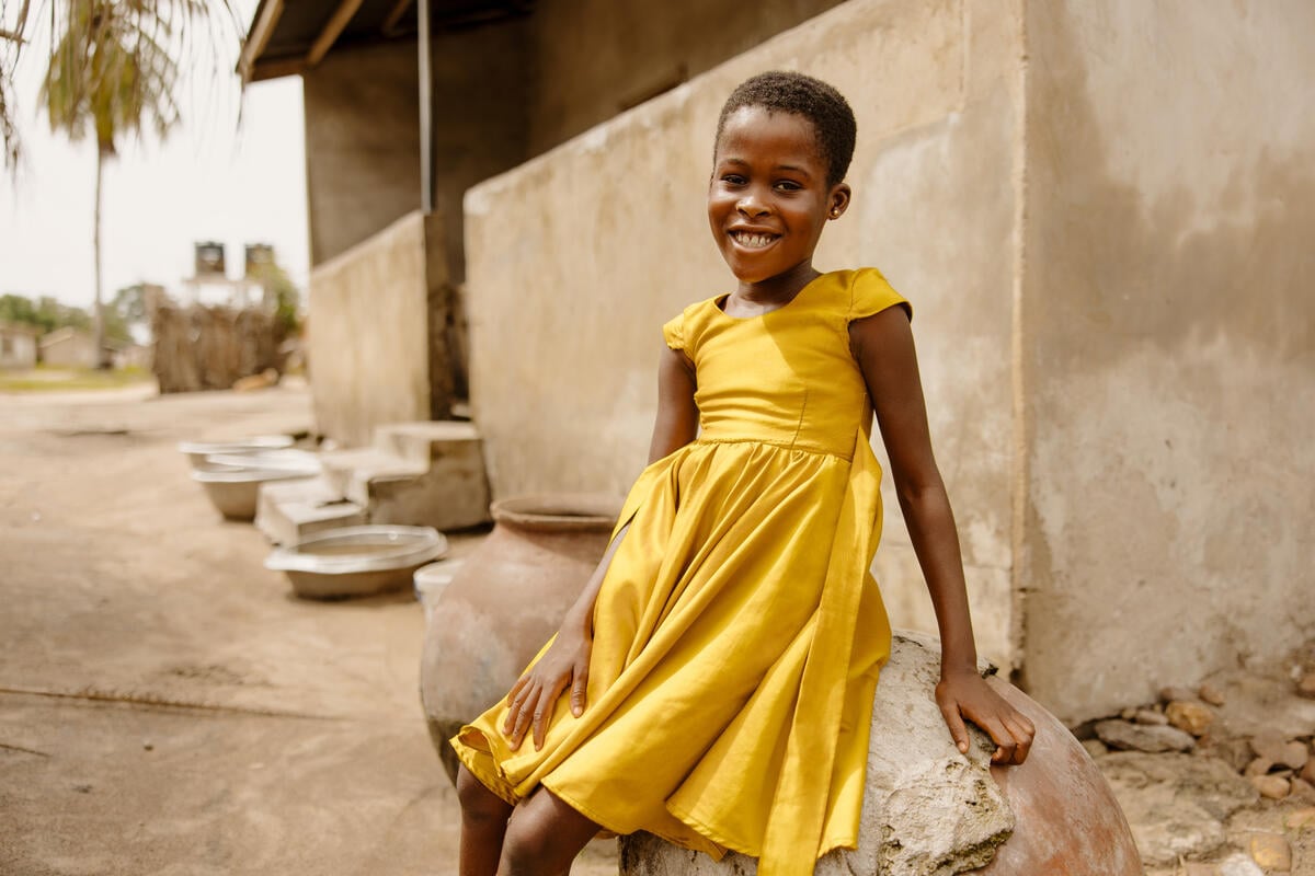 A smiling girl sits on a rock while looking at the camera. There is a building behind her. 