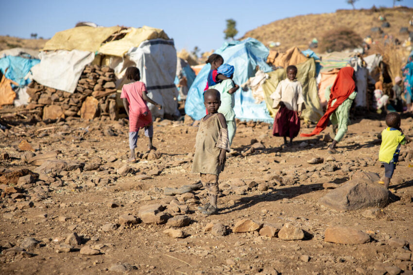 A child stands on dirt amid a displacement camo, looking at the camera.