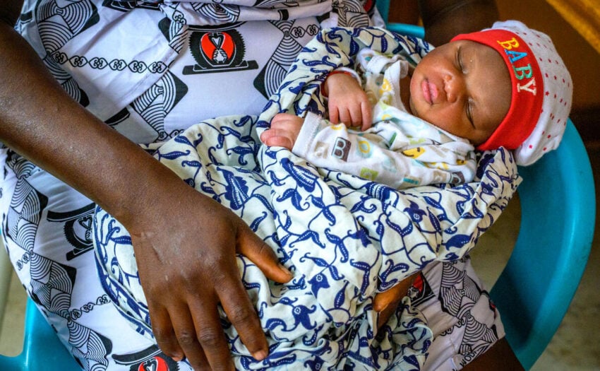 An infant wrapped in a colorful blanket and cap that reads “BABY” sleeps on his mother’s lap, with her hand resting gently on him.
