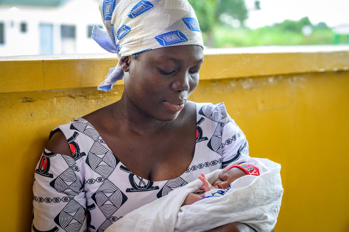 A woman wearing a headscarf holds her newborn baby, swaddled and wearing a colorful knit cap that reads &ldquo;baby.&rdquo;