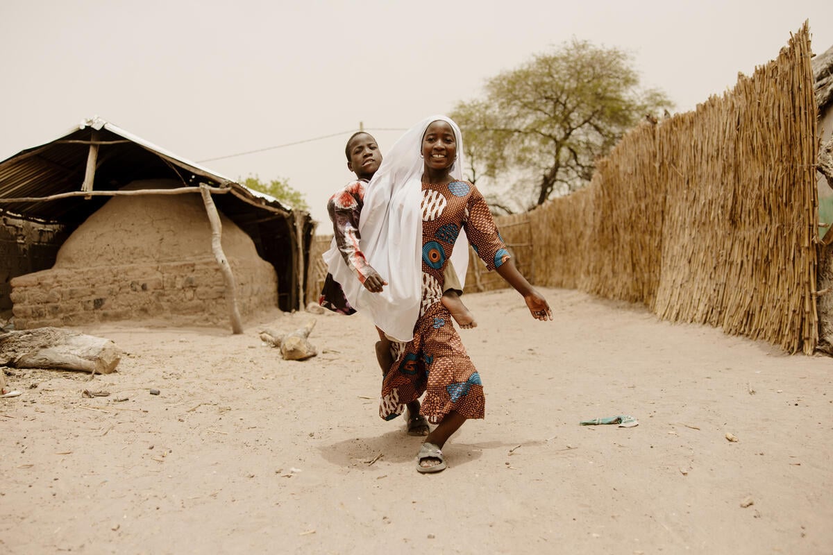 A girl in a long scarf carries a younger boy outdoors on sandy ground in a rural Senegalese community. 