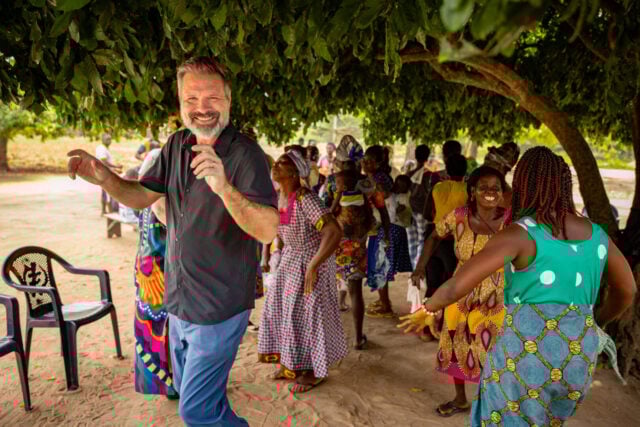 A man and several women dance beneath a tree, wearing colorful clothing, with others gathered nearby