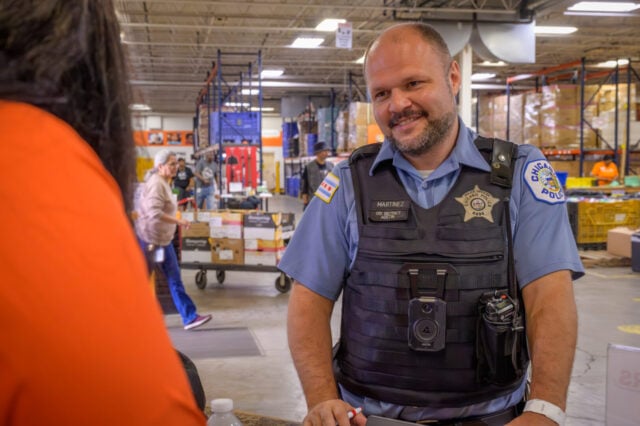 A police officer stands in a warehouse and smiles at a person in an orange shirt.