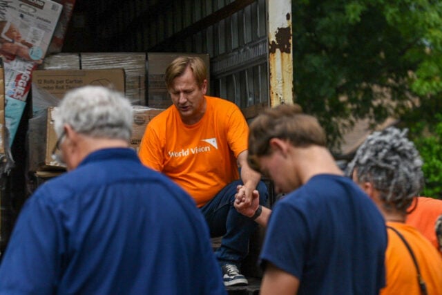 A man in a shirt that reads World Vision sits in the back of the truck holding hands and praying with a group of people.
