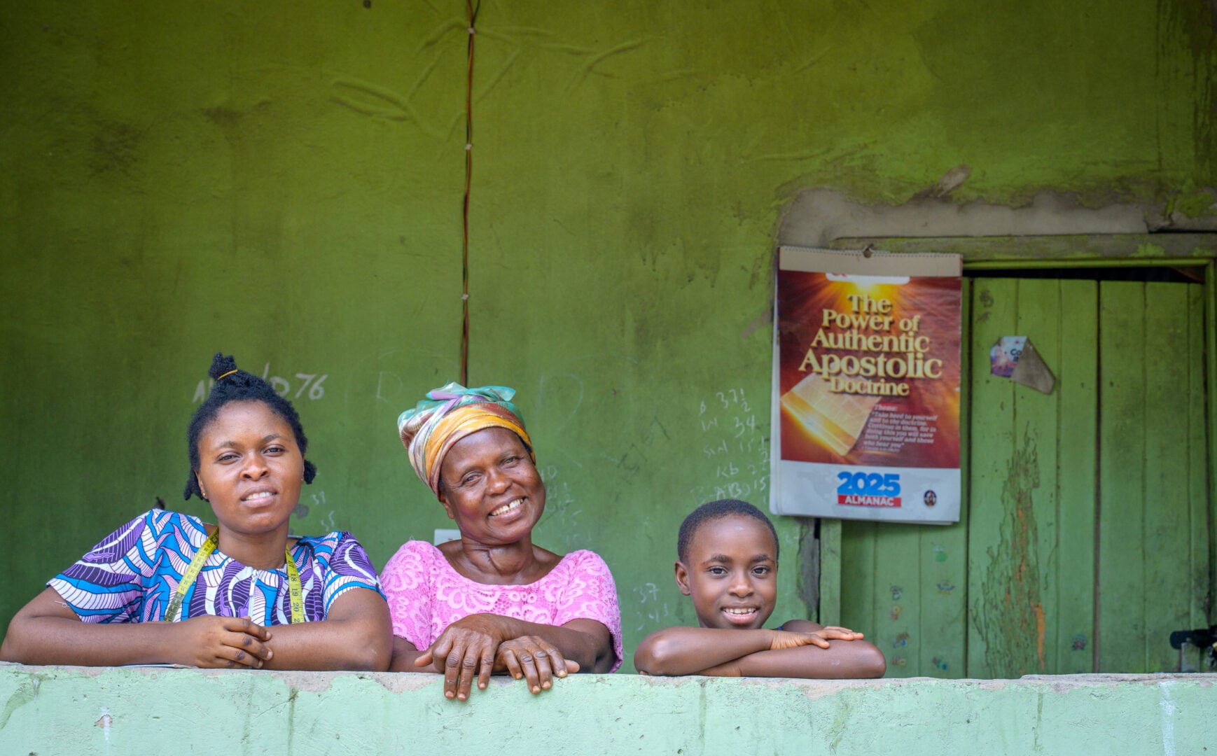 Two women and a girl lean on a low wall as they pose for a photo. Behind them is a wooden door and a calendar hanging on the wall.