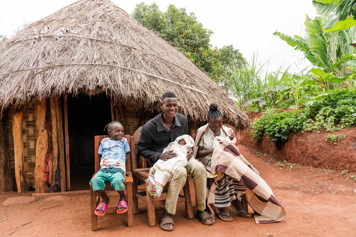 A smiling family sits outside their thatched hut. There is a young child, a father holding a baby, and a mother cradling another baby.